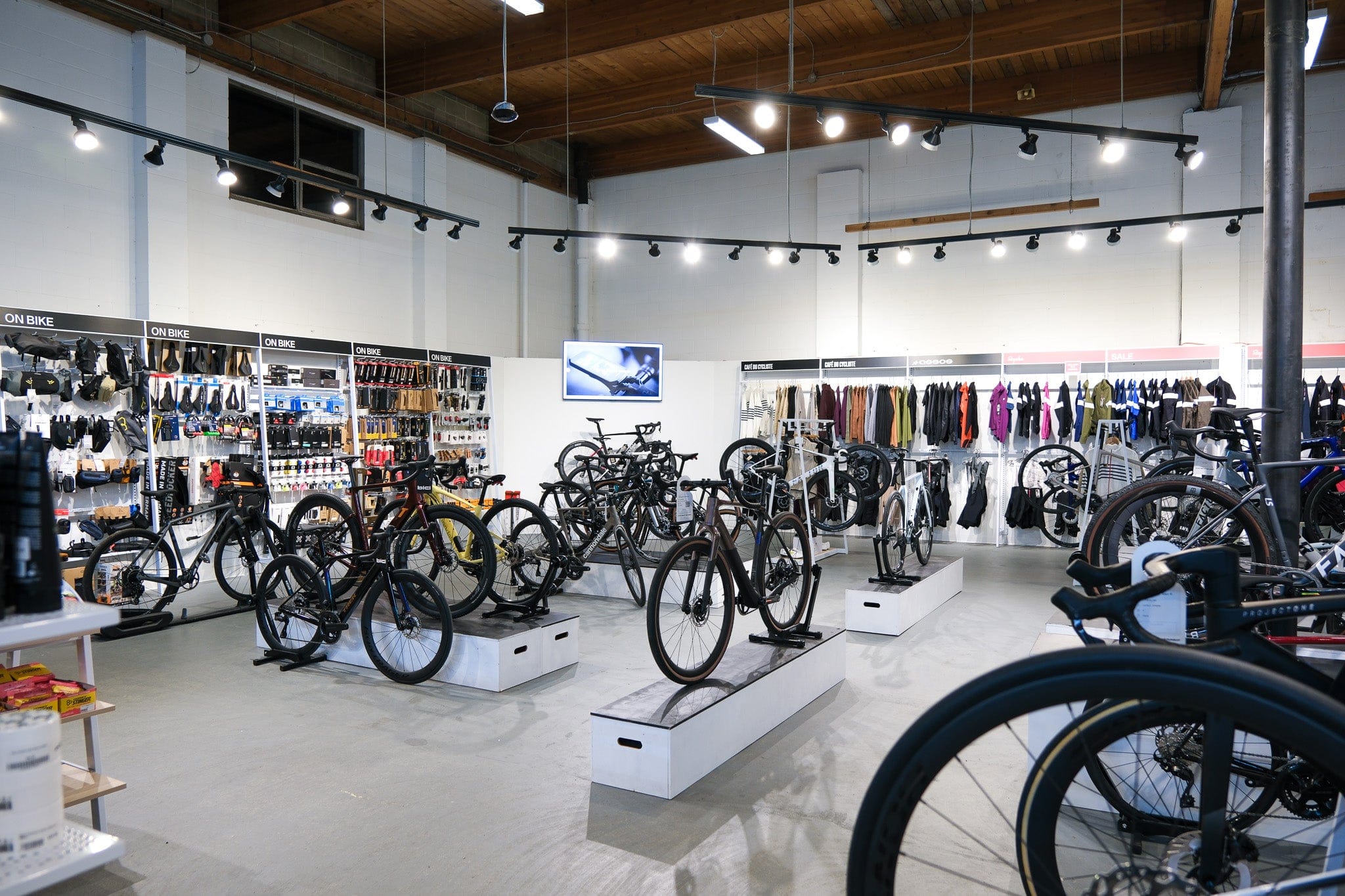 Interior of a sleek bicycle shop with bikes displayed on stands, shelves stocked with helmets, gloves, and cycling tools, and racks showcasing colorful apparel beneath bright ceiling lights.