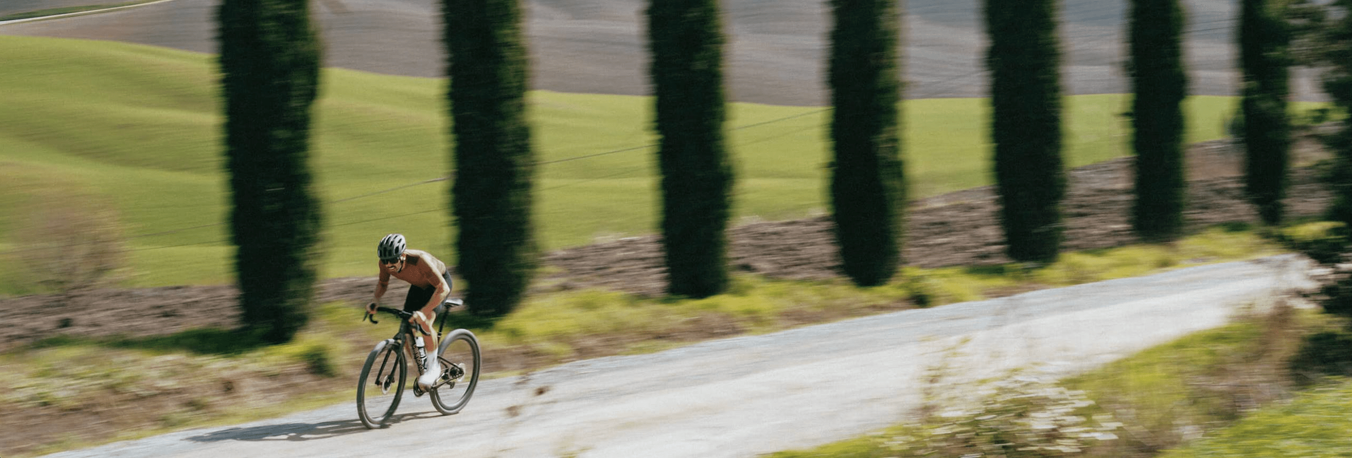 A motion-blurred panoramic shot of a cyclist in a helmet and black shorts riding a bicycle on a gravel road. Behind the cyclist, a line of tall, thin cypress trees stands against a backdrop of rolling green hills.