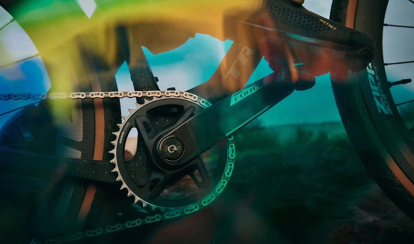 A close-up shot of the drive side of a bicycle, focusing on the crankset, chainring, and chain. The "FORCE" logo is visible on the crank arm. The image has a surreal, artistic filter with a blue and yellow glow, and a faint silhouette of a rider's leg is visible in the background.