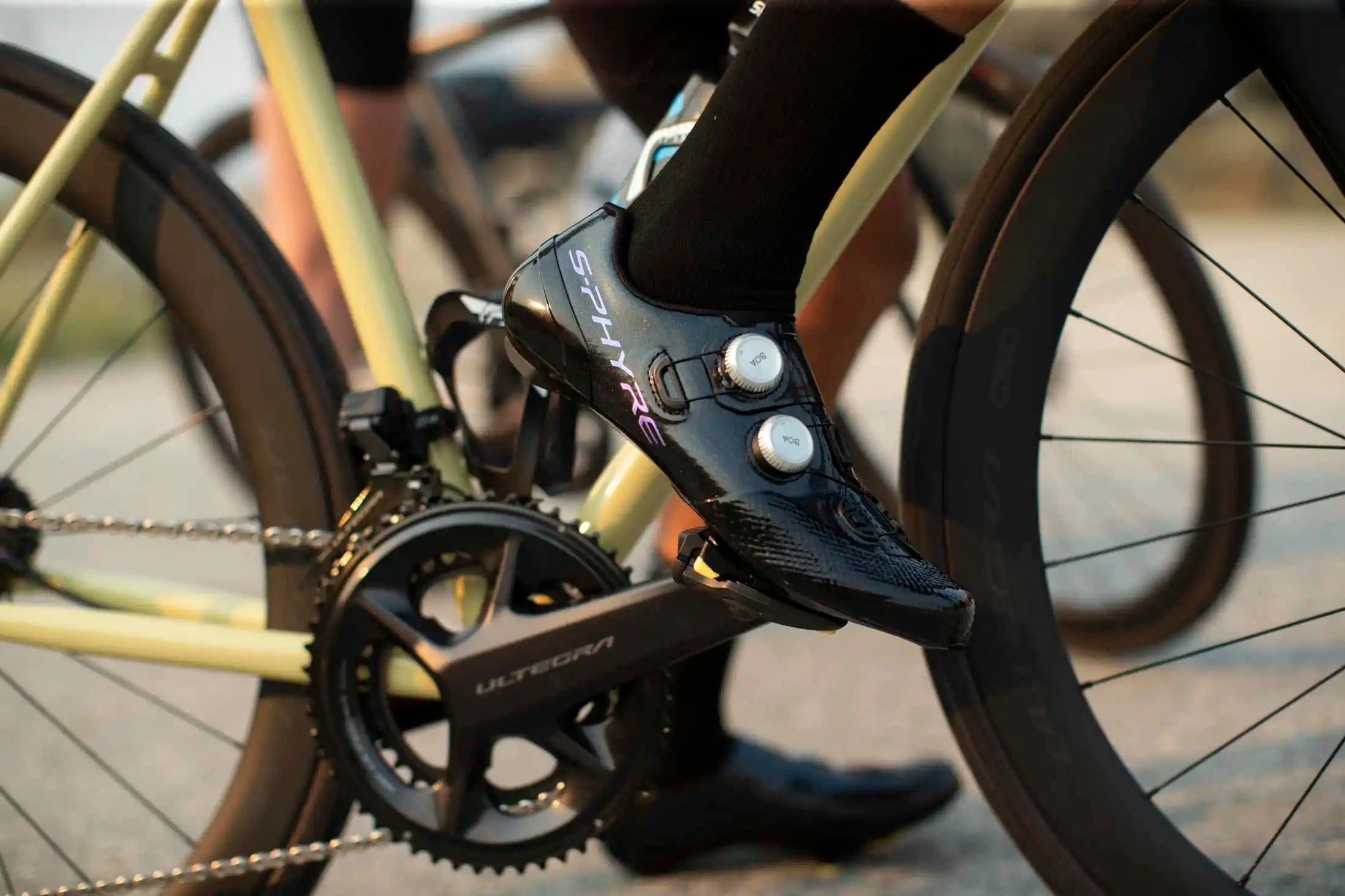 Close-up of a cyclist wearing black Shimano S-Phyre road cycling shoes clipped into an Ultegra pedal on a high-performance road bike with deep-section carbon wheels.