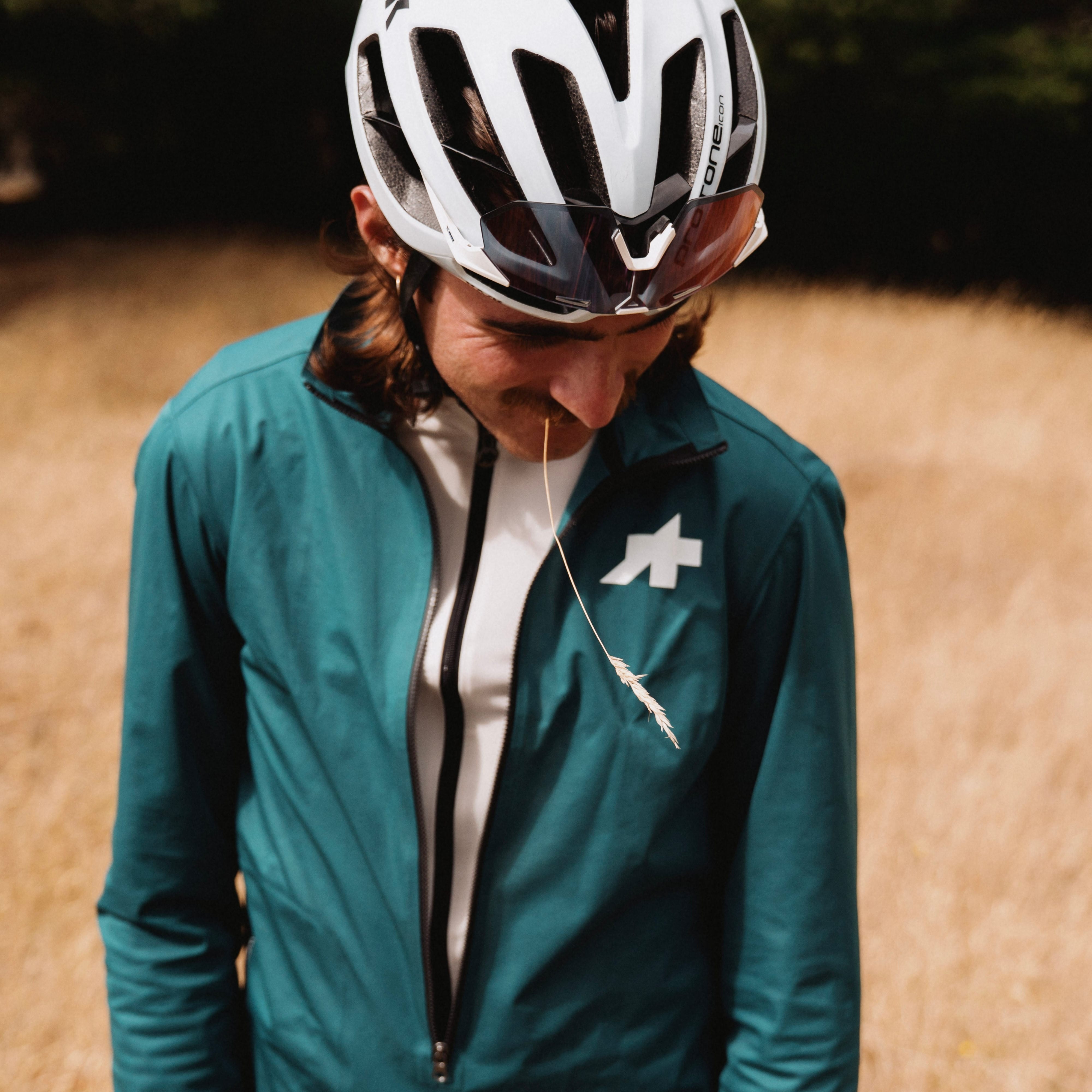 A person outdoors in a dry grassy field wearing a teal cycling jacket over a white top, with a white cycling helmet and dark sunglasses.