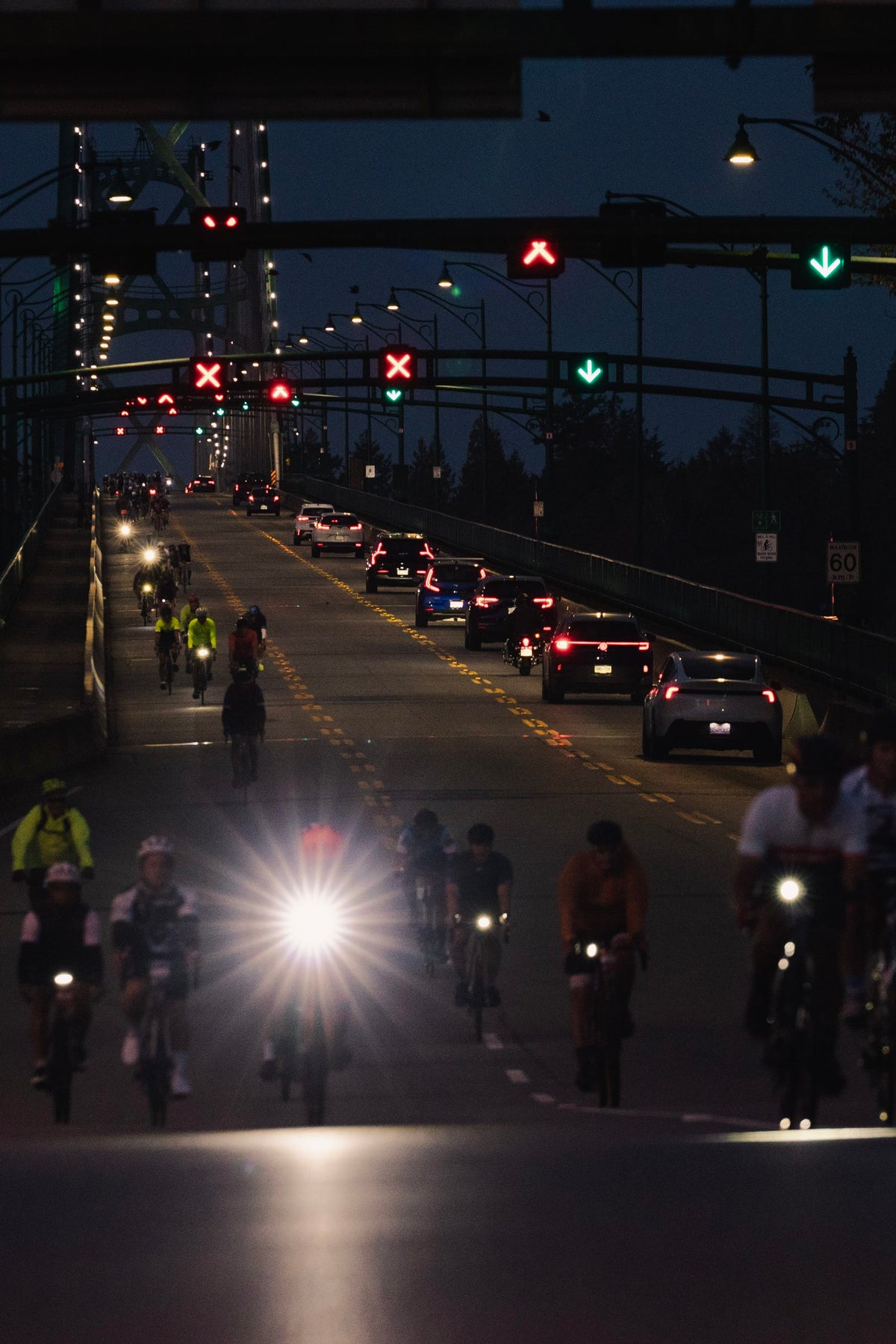 A nighttime street scene with multiple cyclists riding on a bridge or highway, alongside cars with headlights on. Traffic lights and overhead signs are visible in the dark environment.