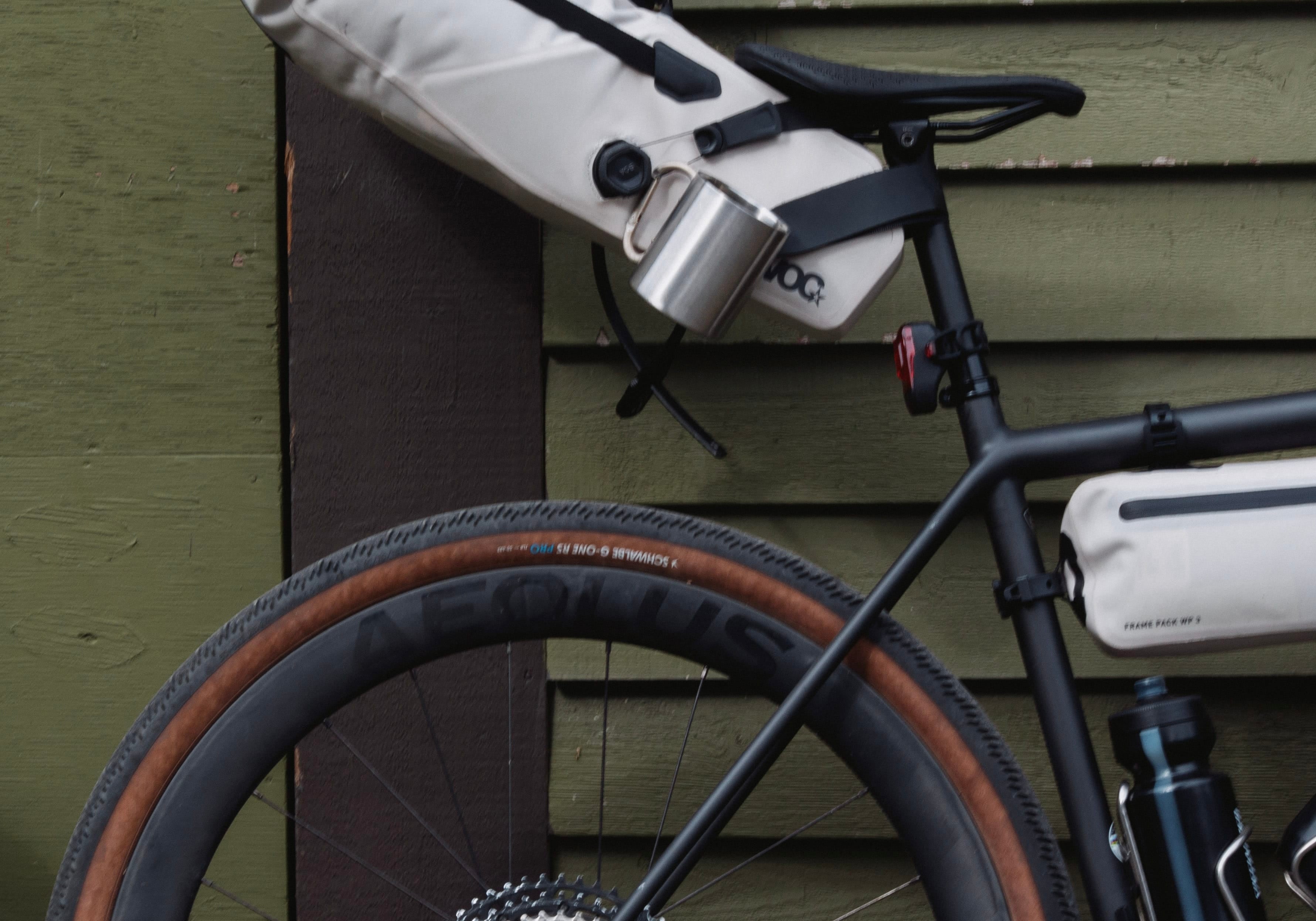 A black bicycle with a white top tube bag and a cream saddle bag leans against an olive green wooden wall. A silver water bottle and a black shoe are attached to the saddle bag.