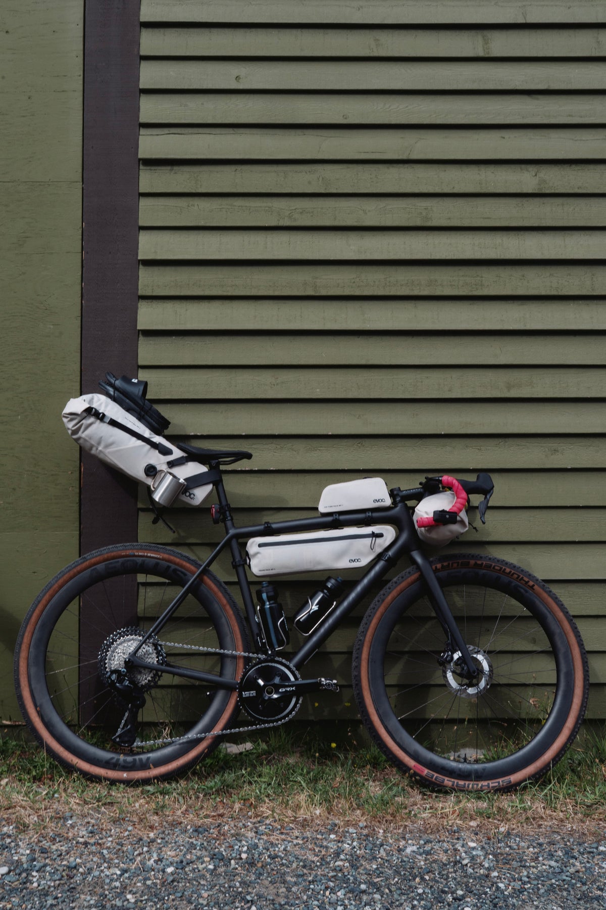 A black gravel bicycle with white and gray pannier bags and a red front bag, parked against an olive green wooden wall.