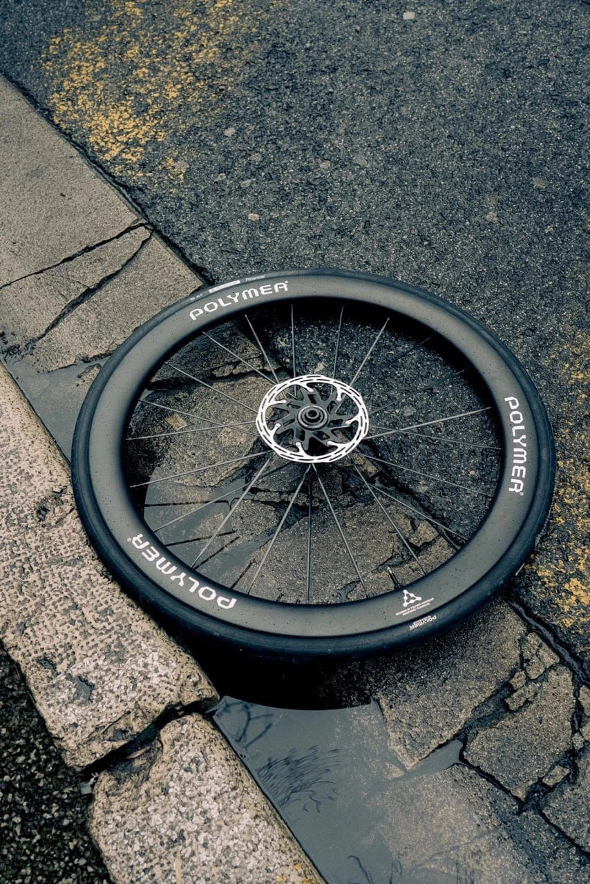 A Polymer Workshop carbon road wheel with disc brake rotor rests on a cracked asphalt curb, partially in a rain puddle. The scene highlights the wheel’s durability and street-tested aesthetic