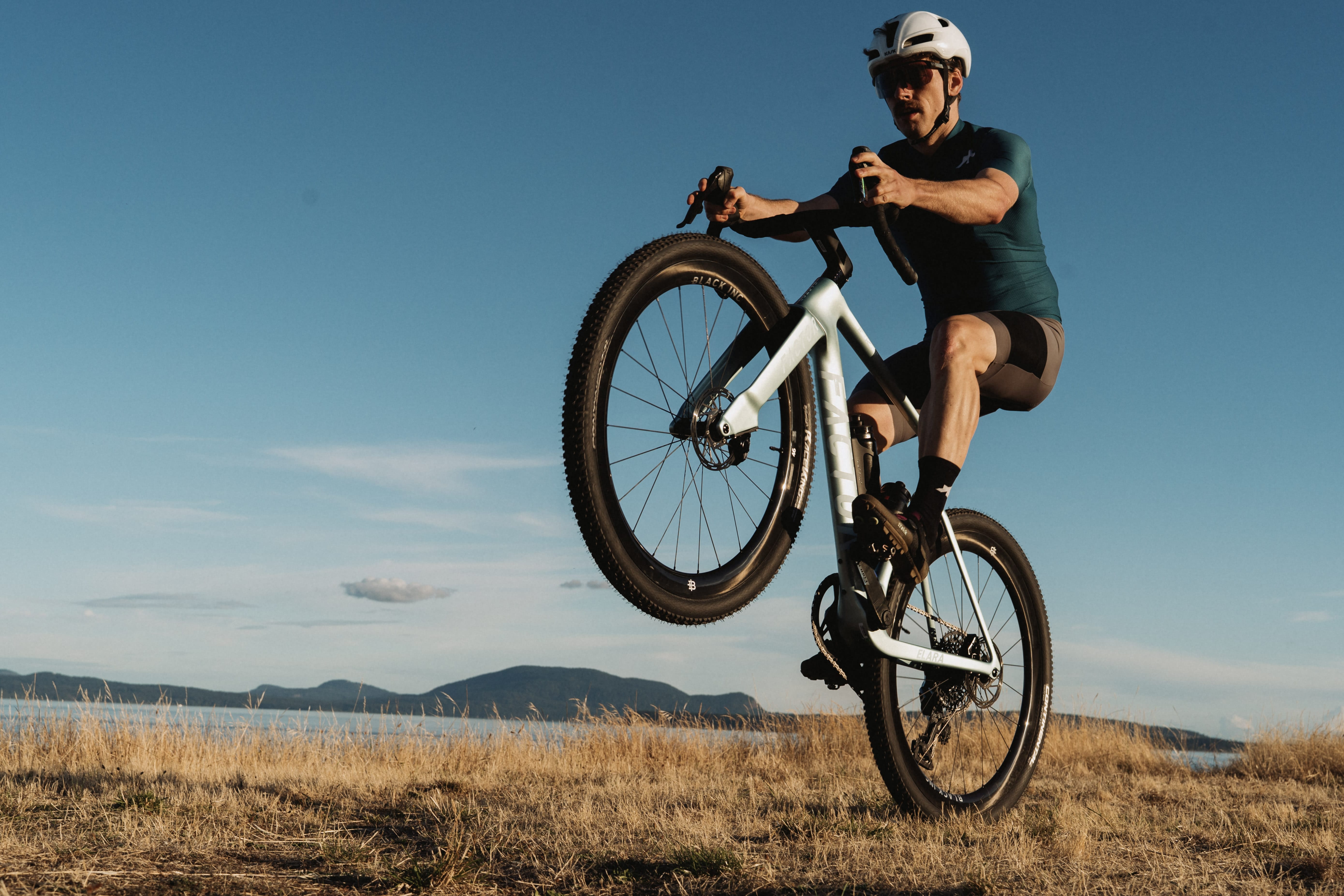A man in cycling gear and a white helmet performs a wheelie on a white gravel bike outdoors on a grassy hill under a clear blue sky.