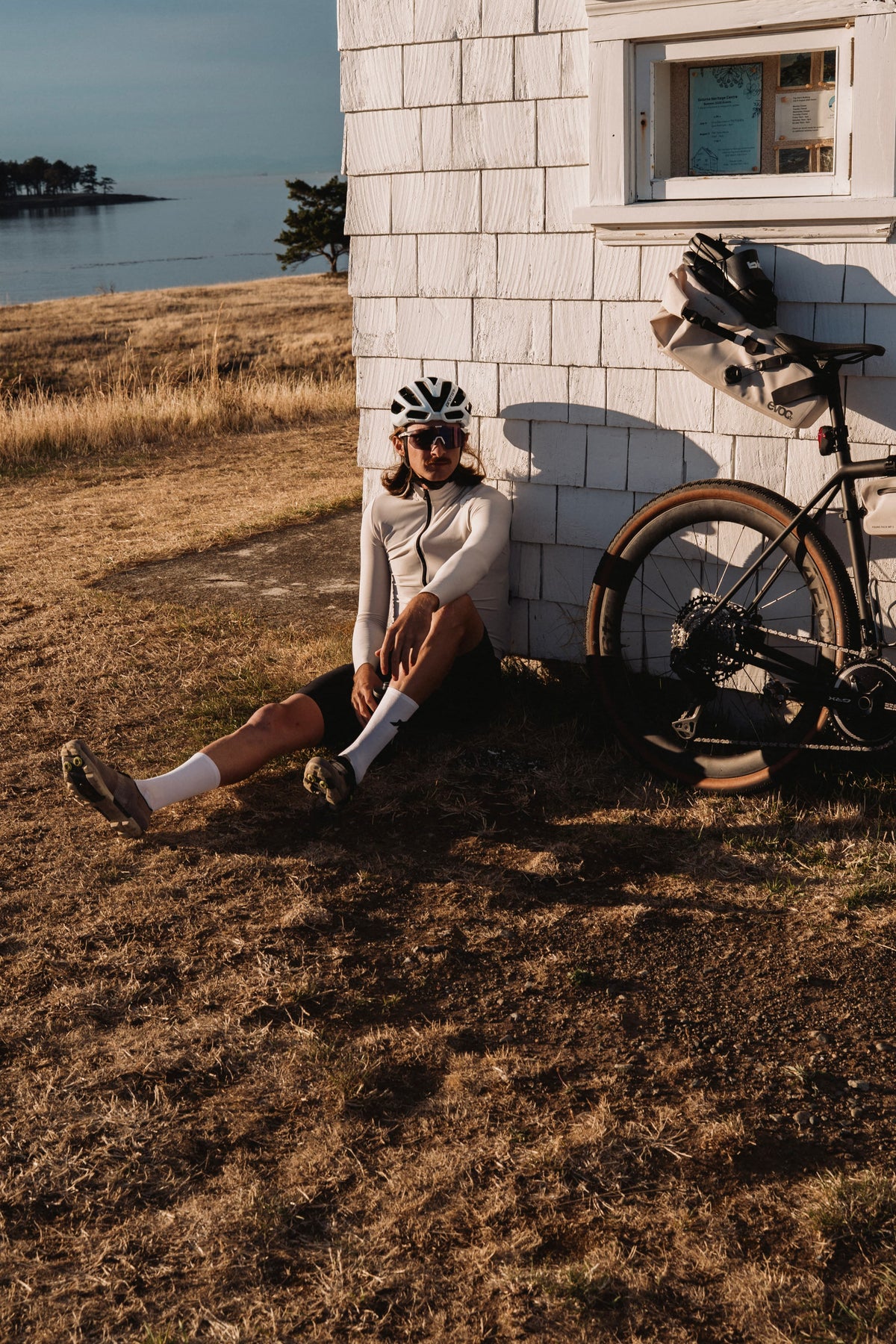 A cyclist wearing a black and white helmet, sunglasses, and a white long-sleeve jersey sits on dry grass next to a white shingled building, with a bicycle and gear leaning against the wall. Water and shoreline are visible in the background.