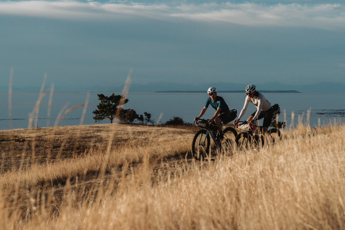 Two cyclists ride along a grassy path overlooking a calm body of water under a cloudy sky, with dry golden grass in the foreground.