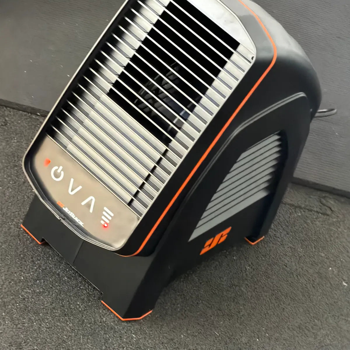 A compact black and orange portable heater or fan with a slotted front grille, photographed on a dark gray paved surface.