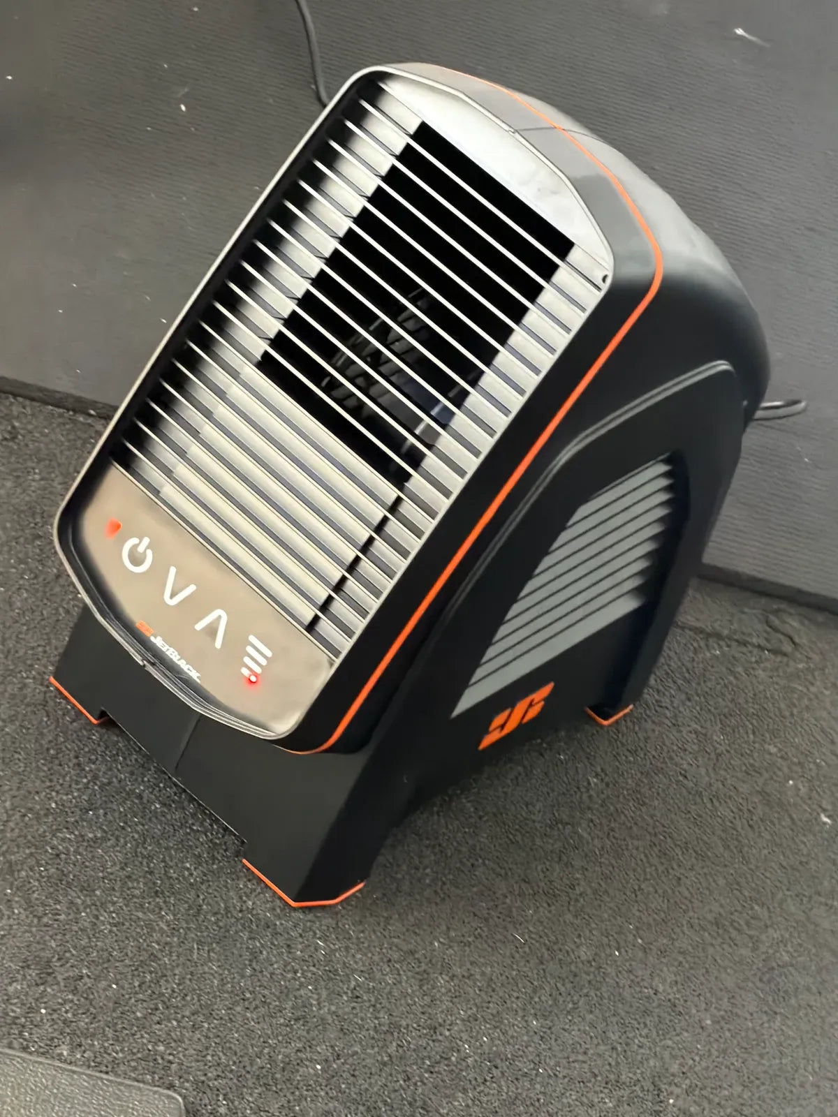 A compact black and orange portable heater or fan with a slotted front grille, photographed on a dark gray paved surface.