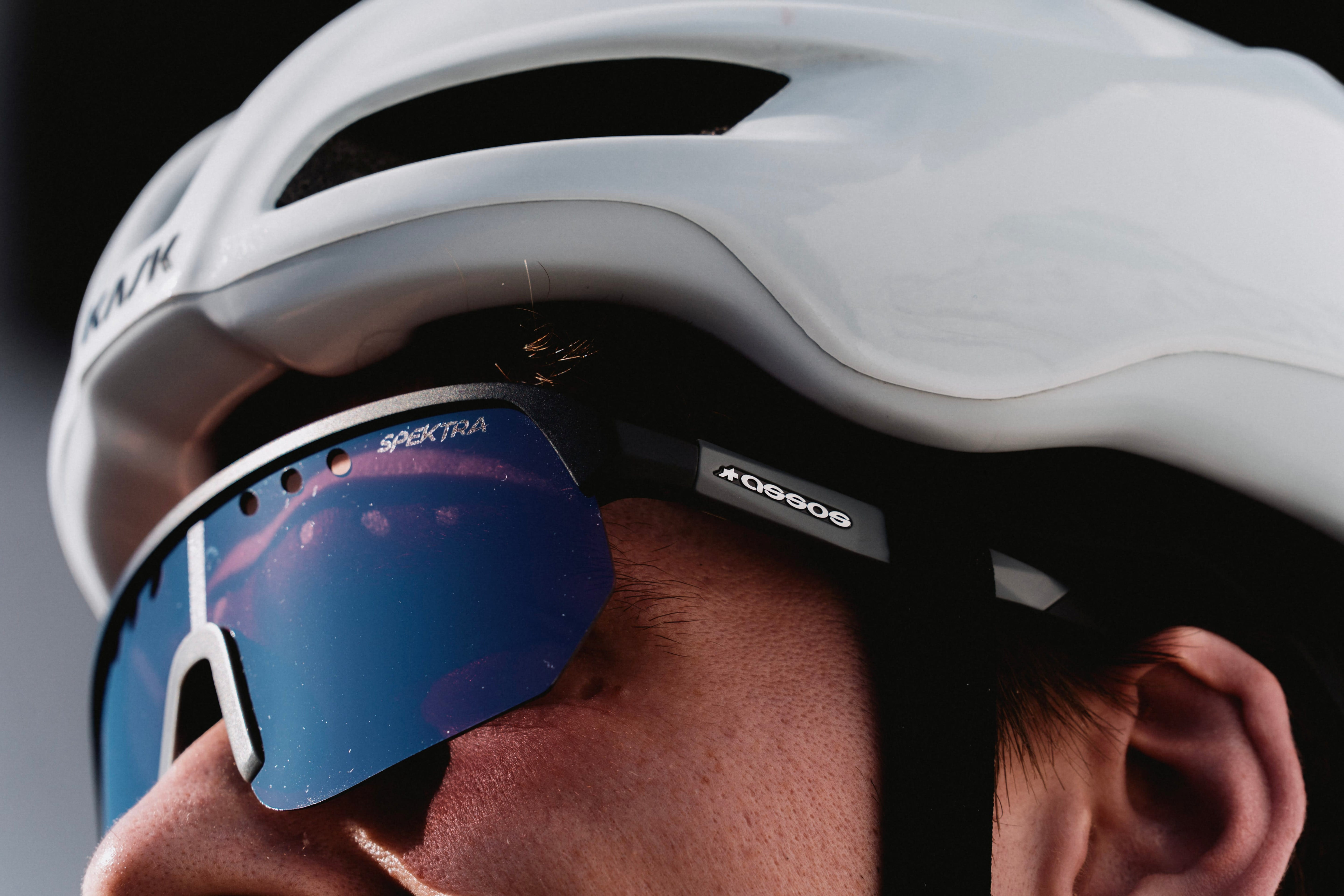 Close-up of a cyclist wearing a white Kask helmet and black sunglasses with blue mirrored lenses. The brand name 'KASK' is visible on the side of the helmet, and 'SPECTRA' is visible on the lens frame.