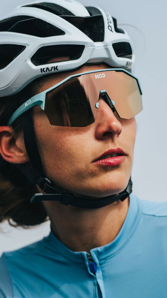 Close-up of a female cyclist wearing KOO mirrored sunglasses and a white Kask helmet, dressed in a light blue cycling jersey