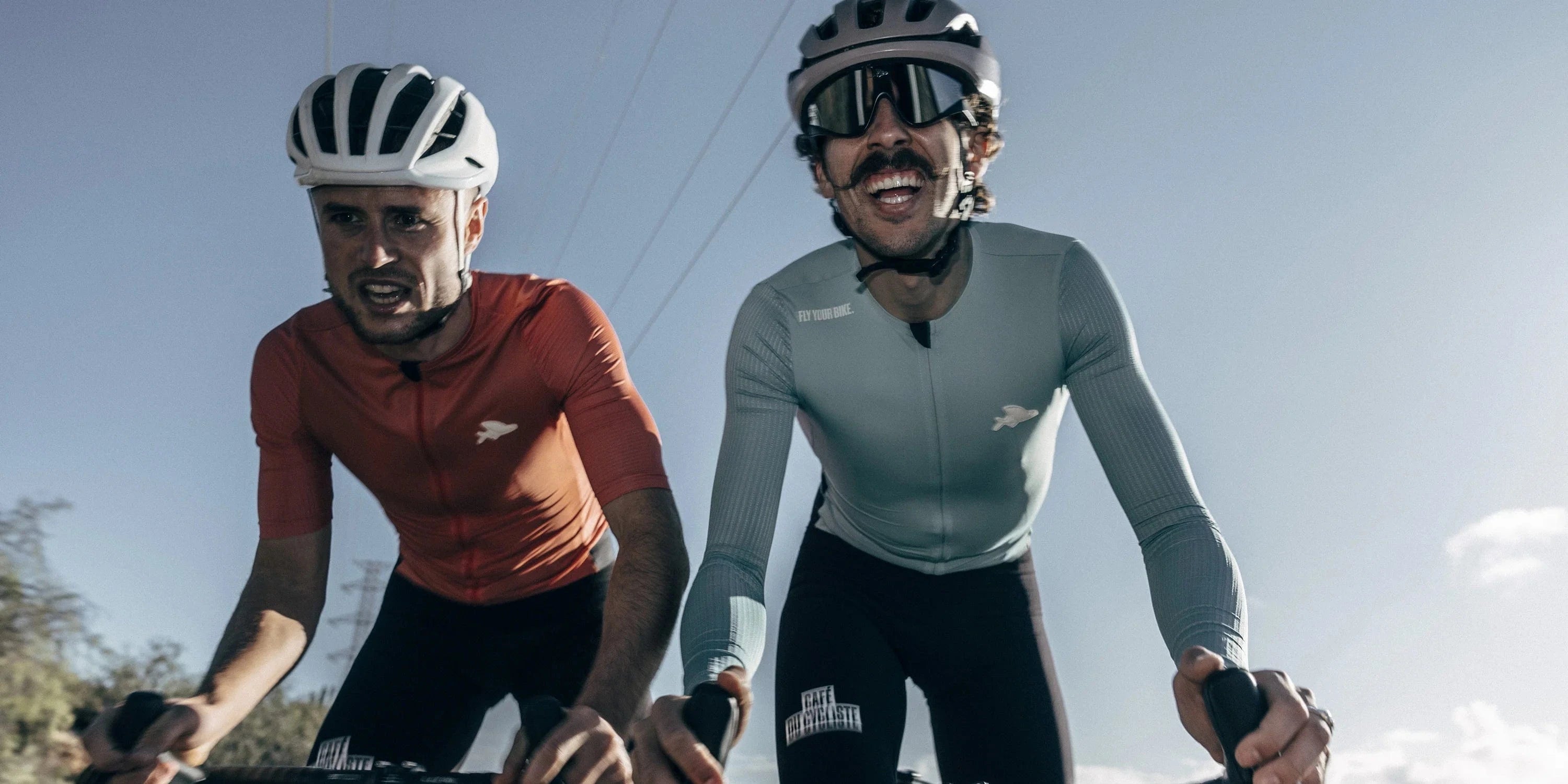 Two cyclists wearing Café du Cycliste jerseys ride side-by-side under a bright blue sky. One wears a red short-sleeve jersey and white helmet, pushing hard. The other, in a pale blue long-sleeve jersey and pink helmet, grins while riding. Both wear Café du Cycliste bibs and appear mid-effort on a sunny climb