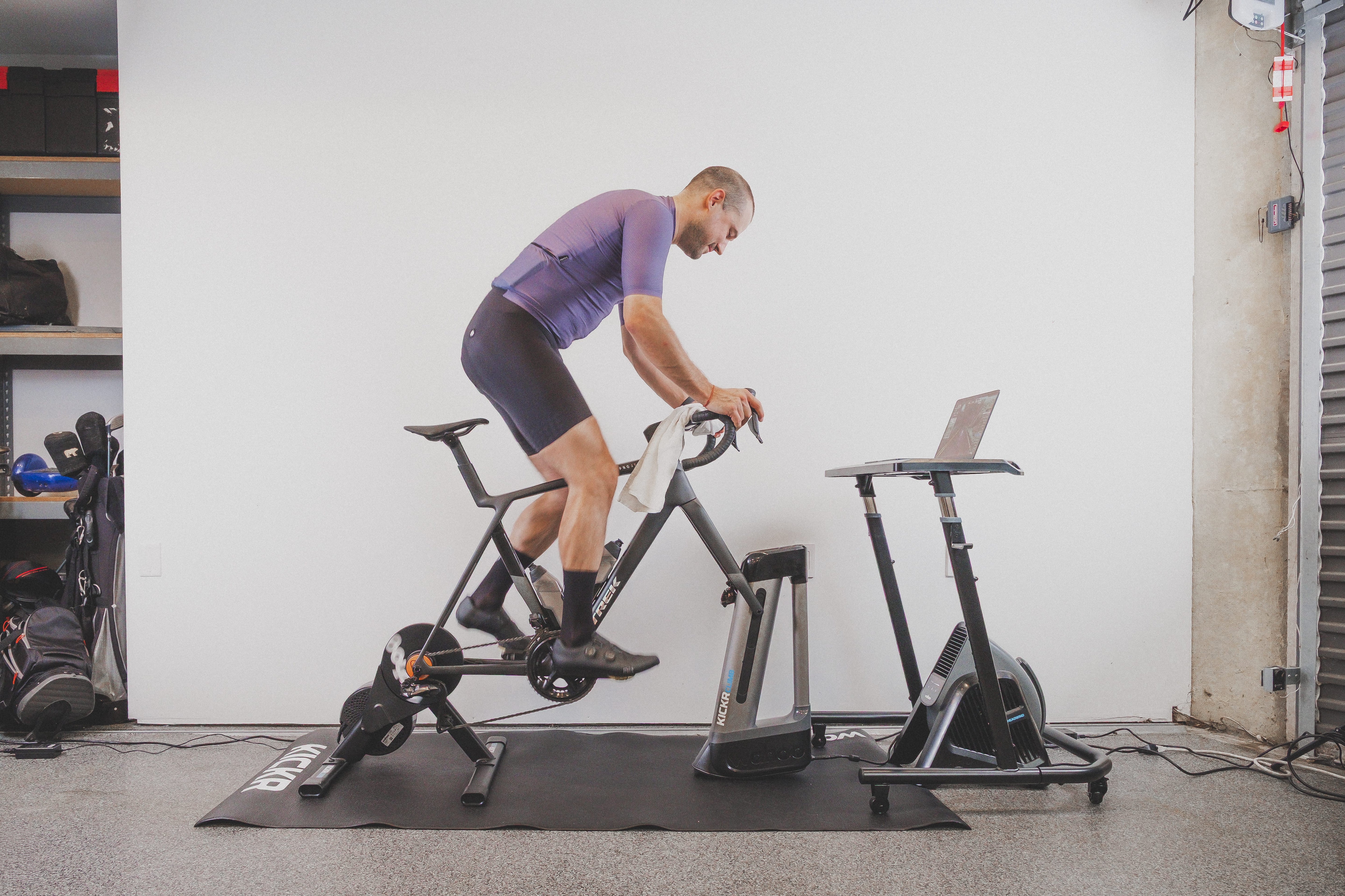 A man in a purple cycling jersey and black shorts rides an indoor stationary bike with an attached crank and a laptop on a stand in a home gym setting.