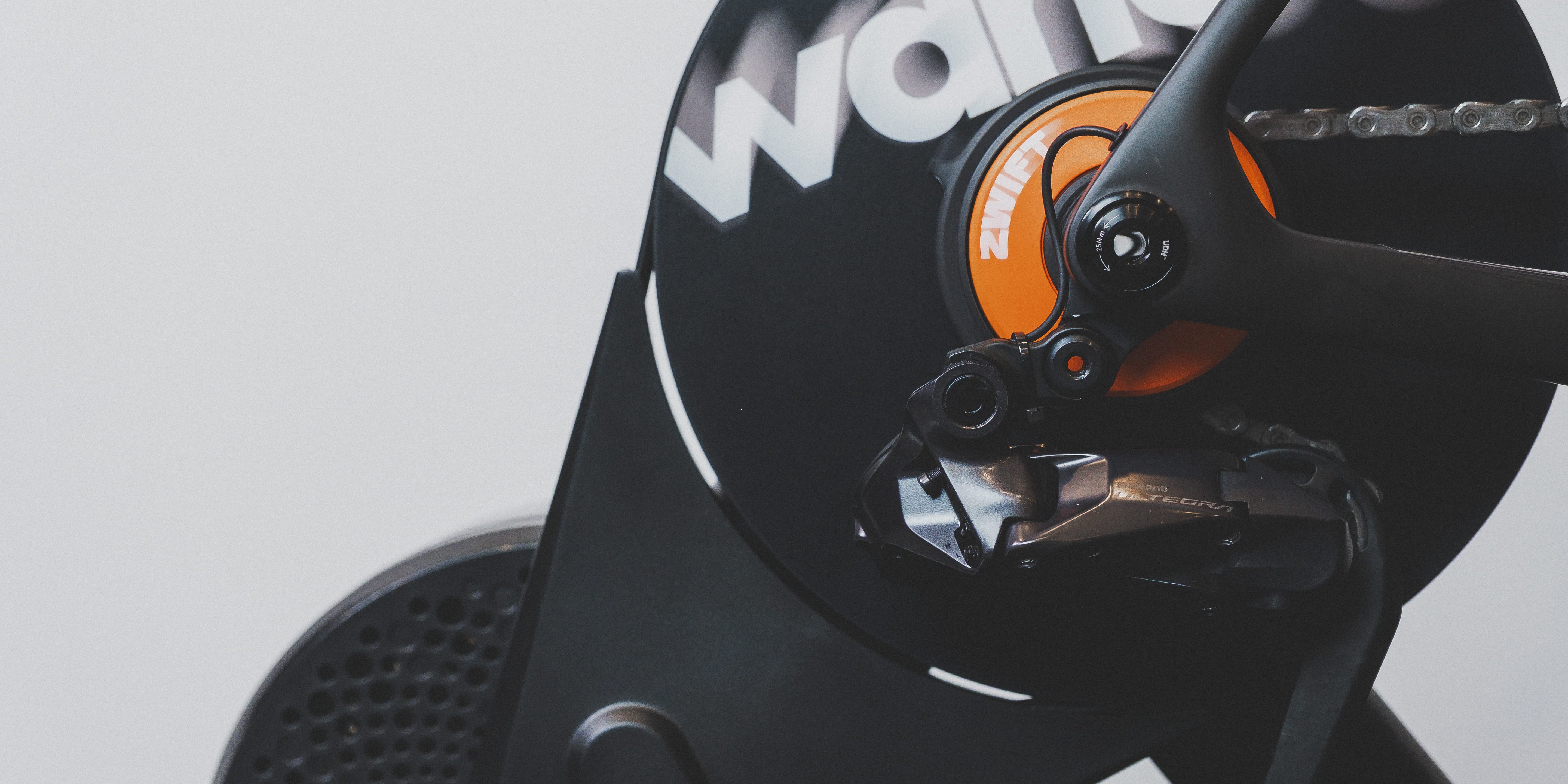 Close-up of a Wahoo indoor cycling computer and chainring assembly, showing the black Wahoo logo, orange accent, and dark metallic components against a light gray background.