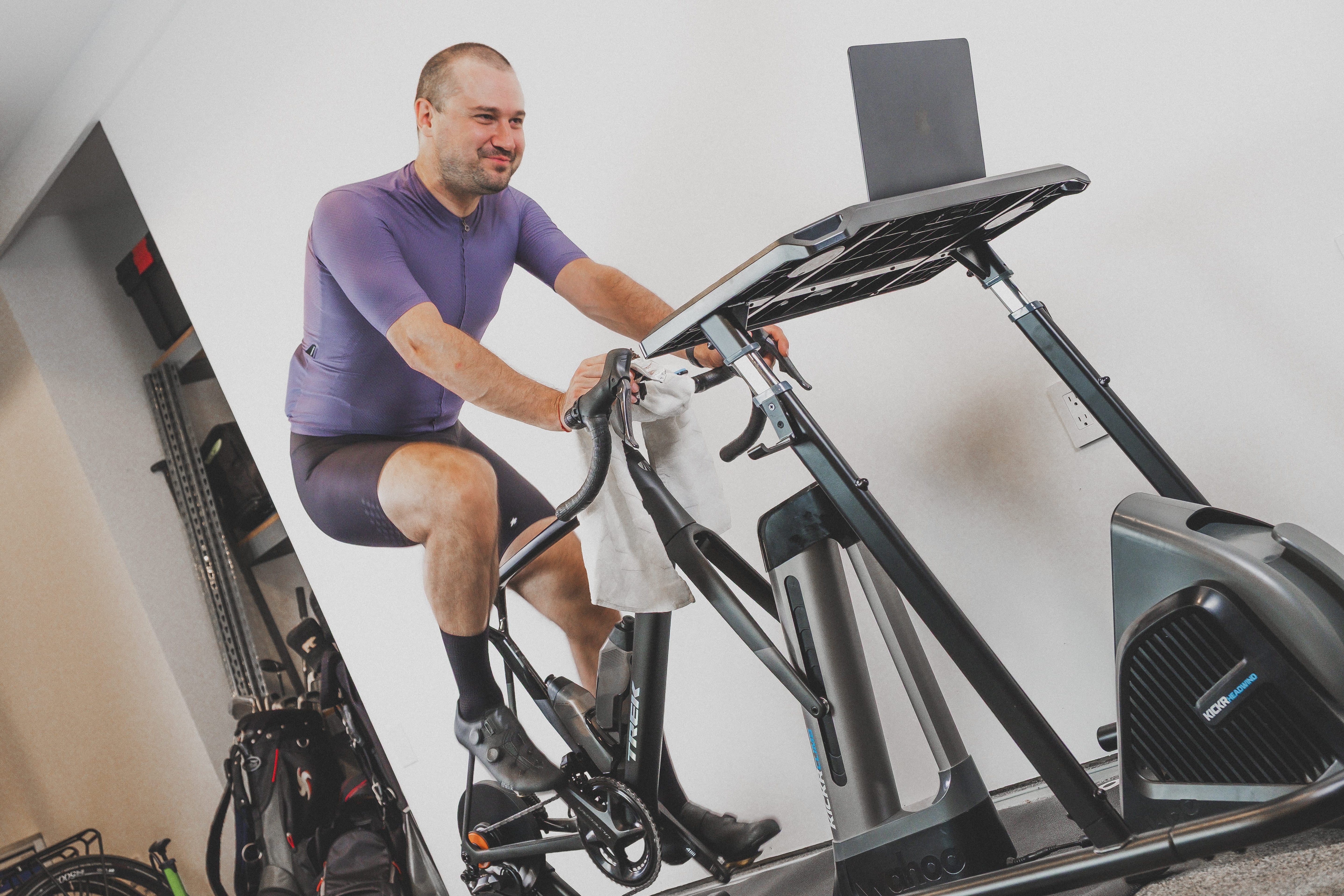 A man wearing a purple cycling jersey and black shorts rides a black indoor stationary bike with a built-in screen and handlebar in a home gym setting.
