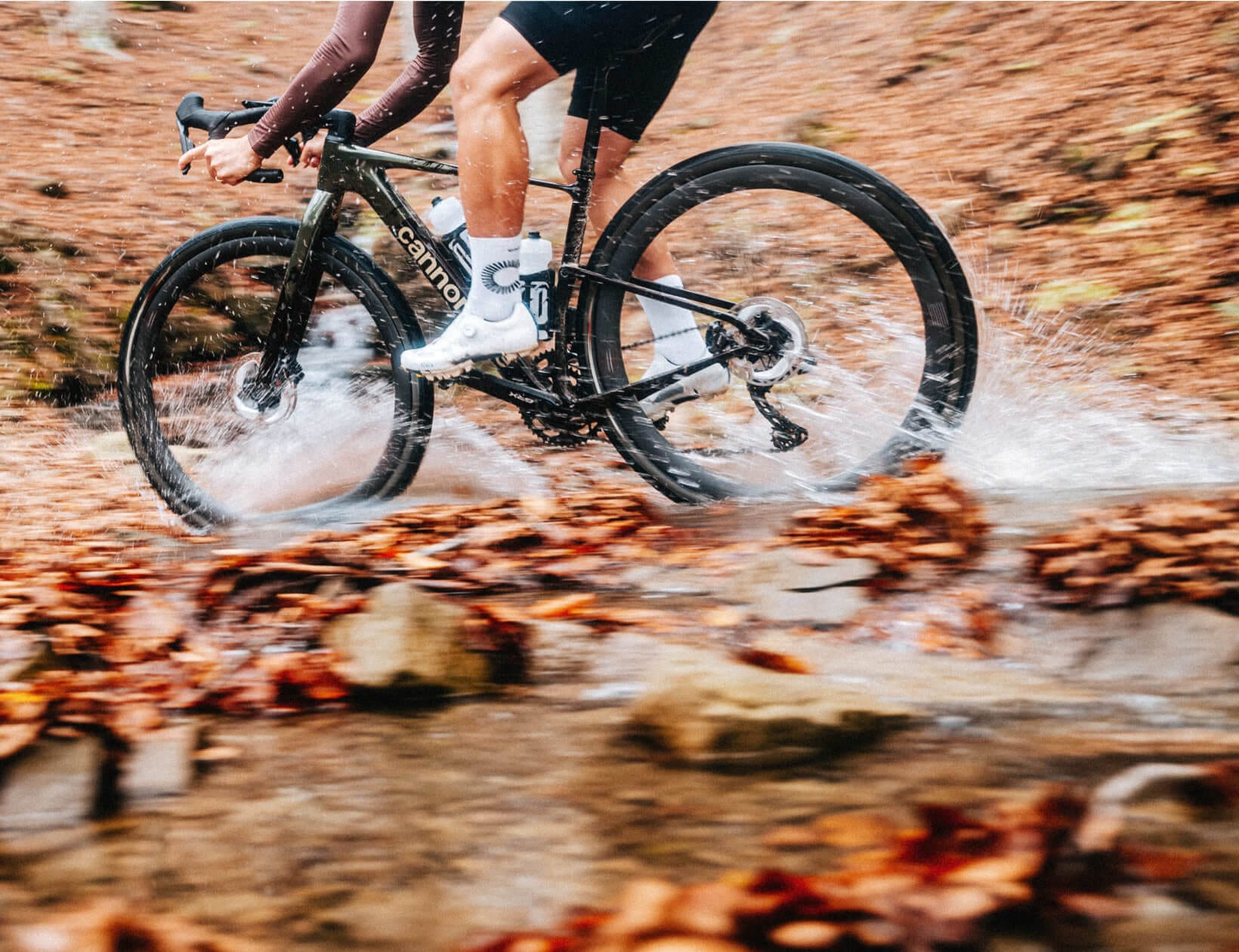 A cyclist rides a Cannondale Topstone through a shallow stream on a forest trail, splashing water and fallen leaves. The bike features disc brakes, deep-section wheels, and white cycling shoes with tall logo socks