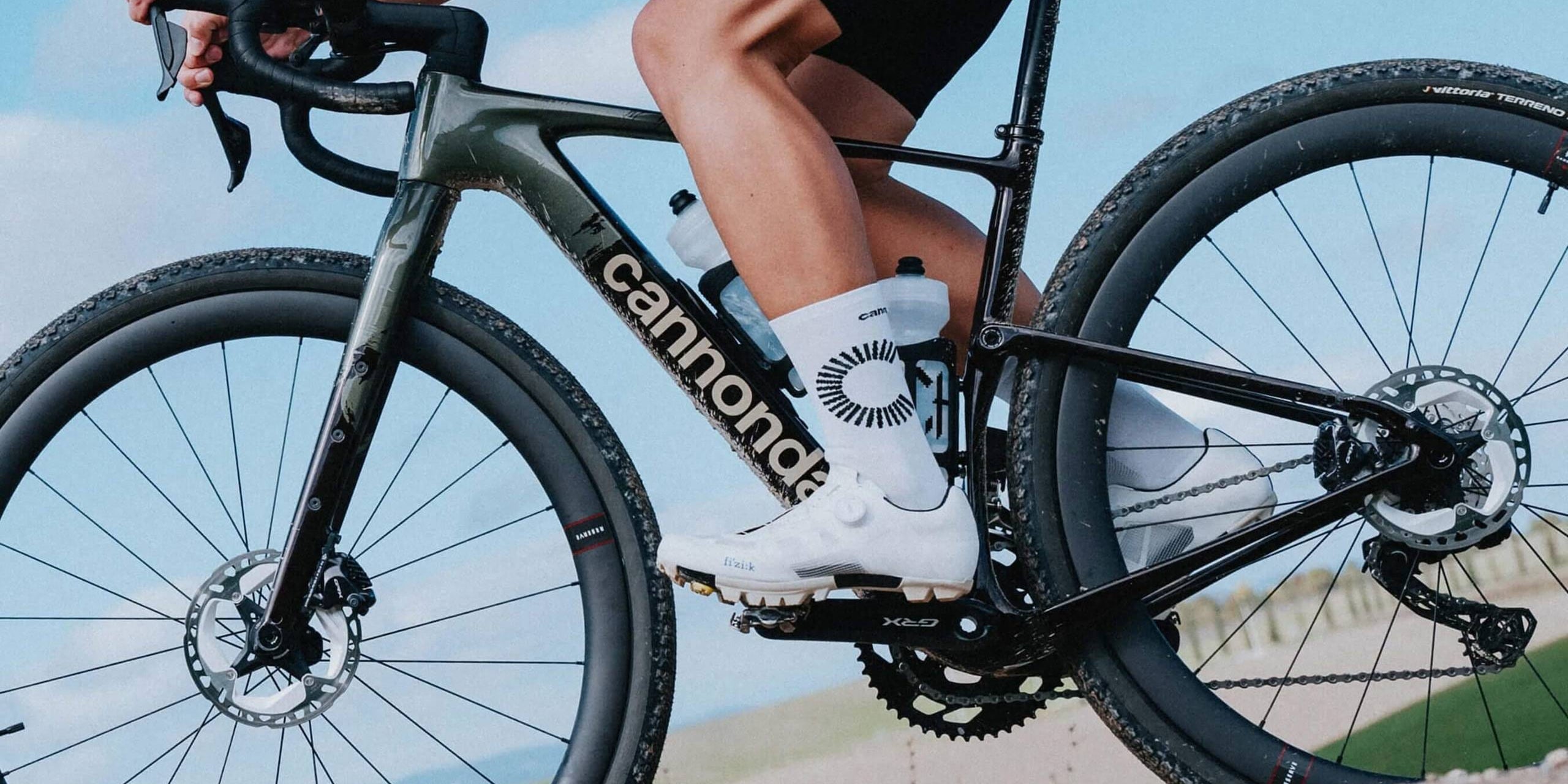 Close-up side view of a rider on a Cannondale Topstone gravel bike, showcasing carbon frame, Vittoria Terreno tires, and SRAM XPLR drivetrain. Rider wears white cycling shoes and tall white socks with black graphic detailing