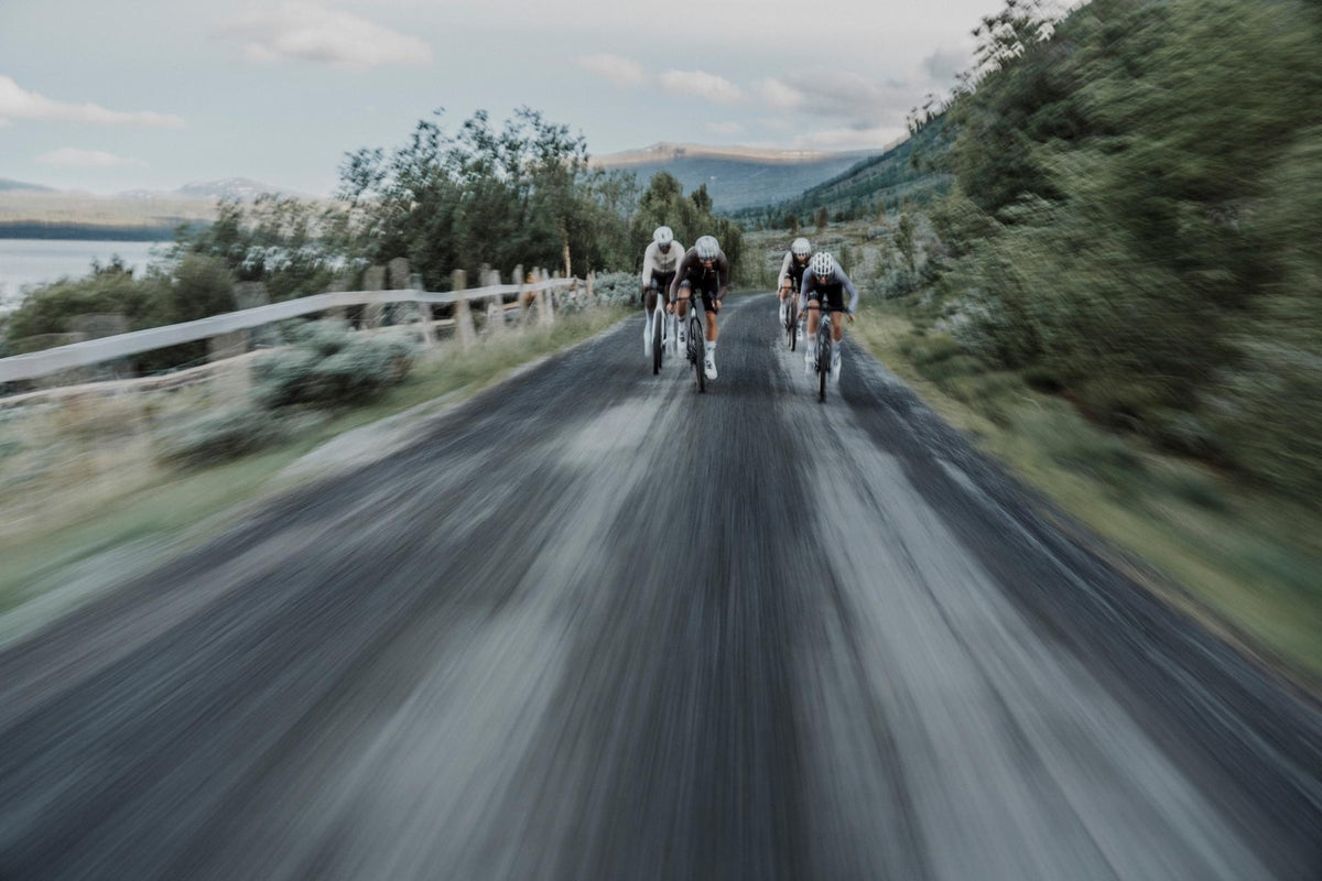 Three cyclists ride away on a paved country road with blurred green trees, a fence, and distant hills under a cloudy sky.