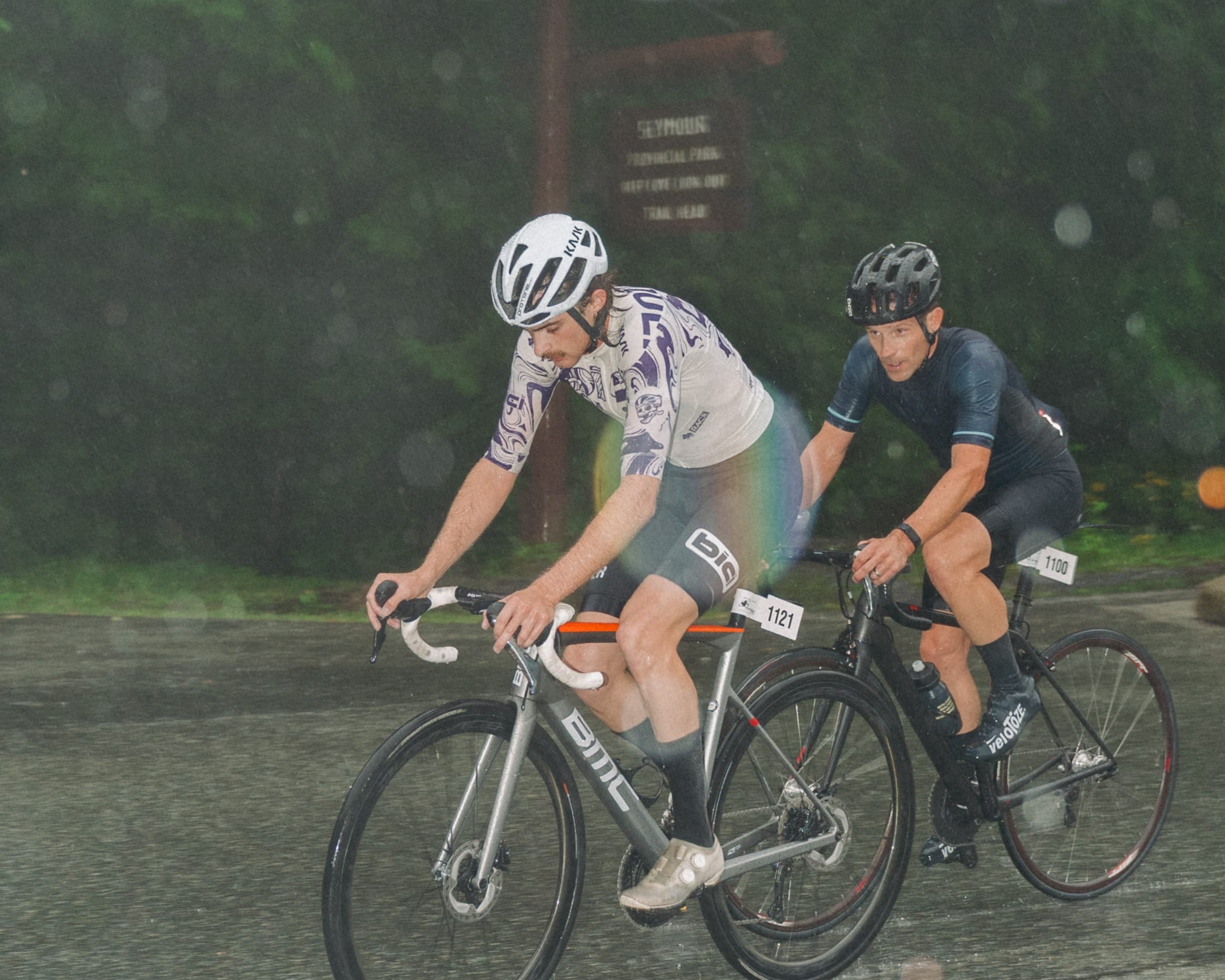 Two cyclists riding road bikes on a wet paved path in the rain, with blurred trees in the background. One cyclist wears a white and gray cycling jersey and shorts, and the other wears a dark navy jersey and shorts.
