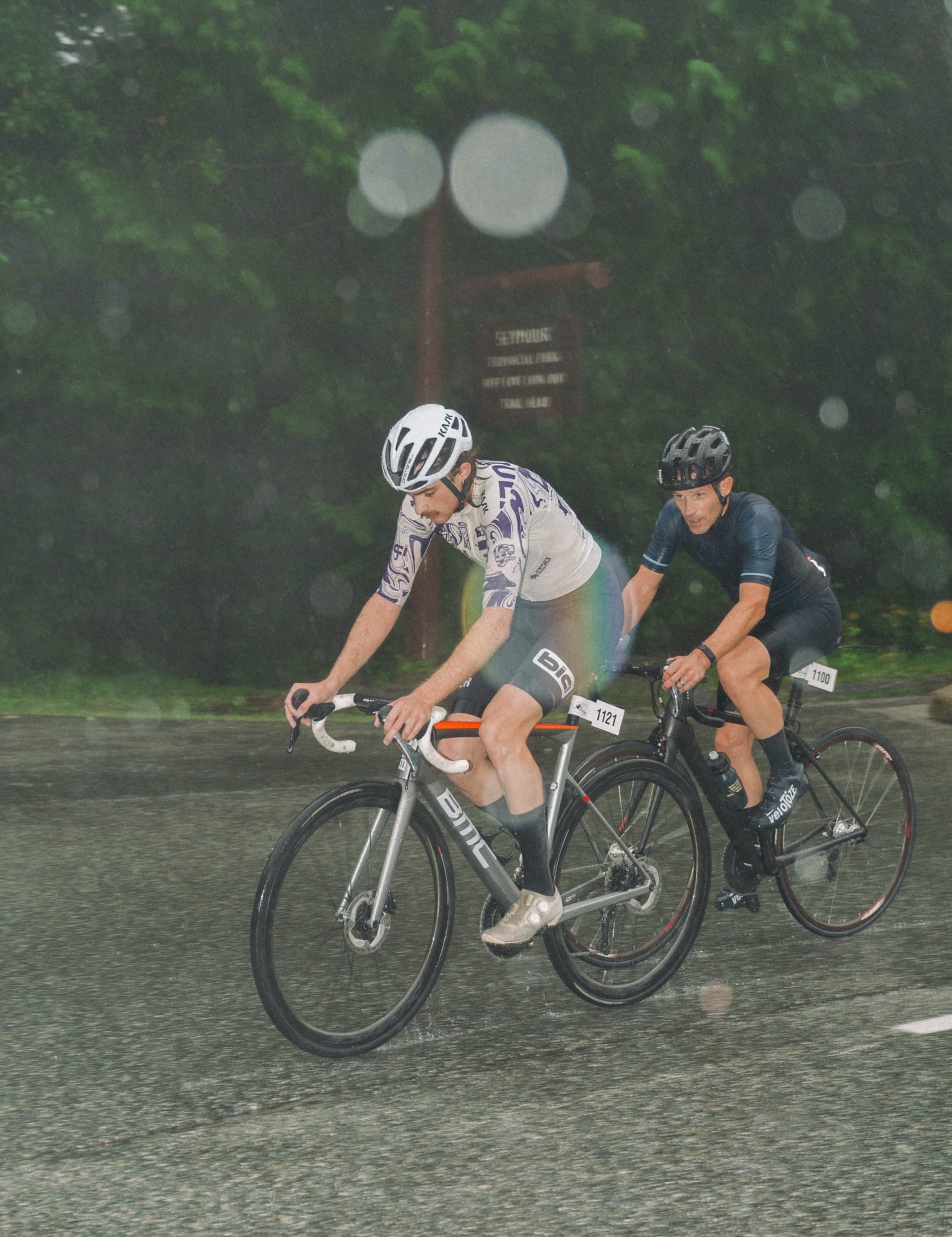 Two cyclists riding road bikes on a wet paved path in the rain, with blurred trees in the background. One cyclist wears a white and gray cycling jersey and shorts, and the other wears a dark navy jersey and shorts.