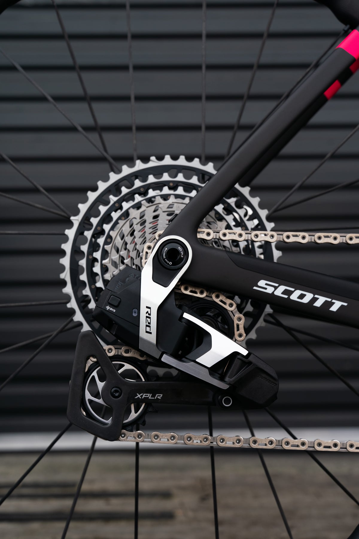 Close-up of a Scott bicycle rear derailleur and cassette area, showing black and silver components with the Scott brand name on the frame, set against a dark metal wall background.