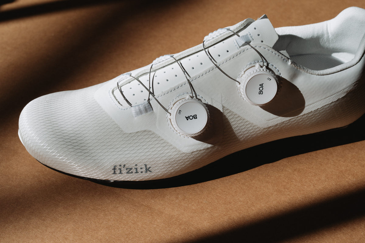 A close-up of a white road cycling shoe with a BOA dial closure system, resting on a brown textured surface with dramatic diagonal shadows.