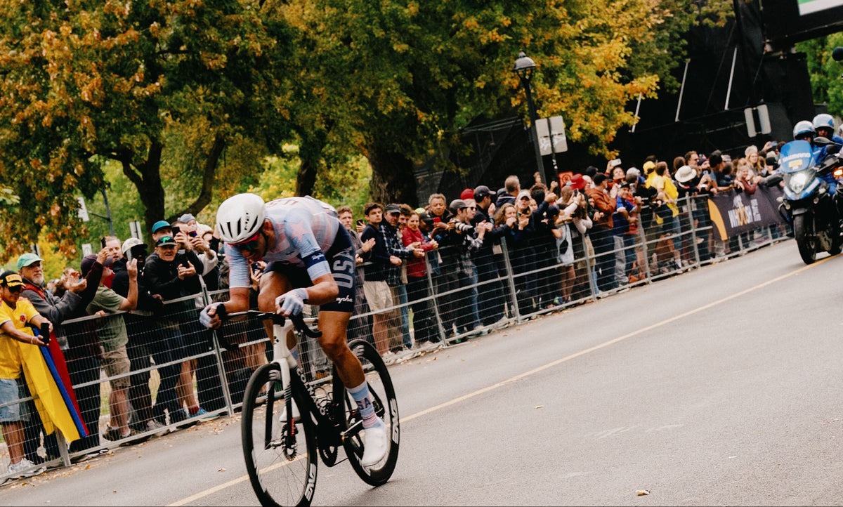 A cyclist in a light blue and white jersey rides a road bike down a city street during a race, with spectators behind metal barriers and trees with autumn foliage in the background.