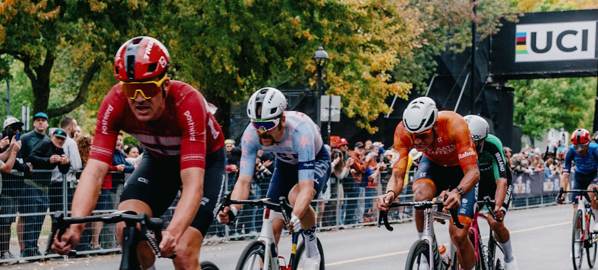 A group of professional road cyclists racing on a city street during a race, with spectators and trees in the background. The cyclists wear colorful jerseys and helmets, riding road bikes with visible UCI branding.