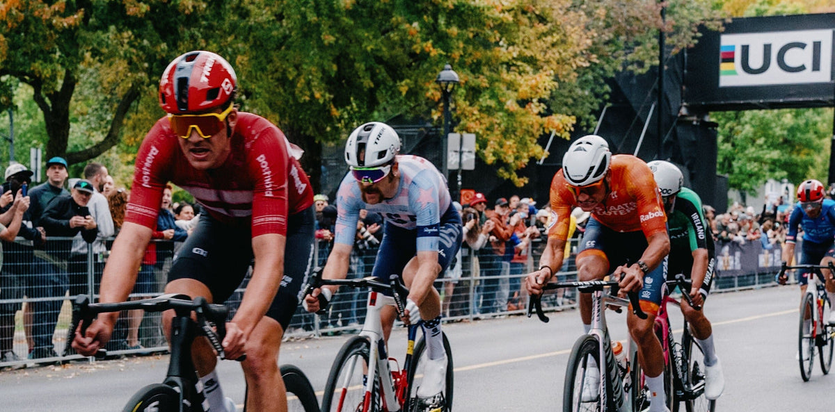 A group of professional road cyclists racing on a city street during a race, with spectators and trees in the background. The cyclists wear colorful jerseys and helmets, riding road bikes with visible UCI branding.