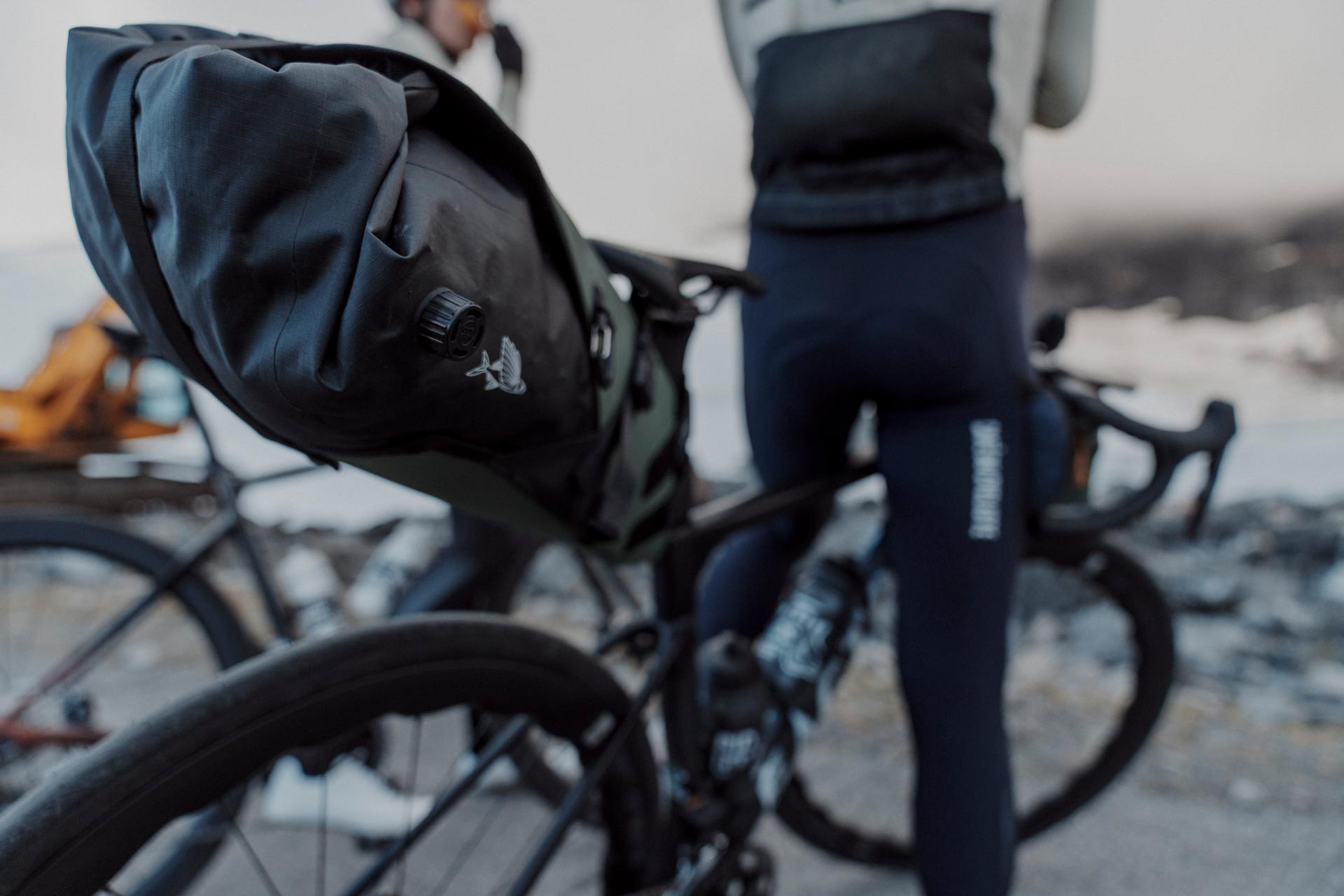 A black waterproof bicycle pannier bag mounted on the rear rack of a bicycle, shown outdoors with a cyclist in dark cycling tights standing nearby.