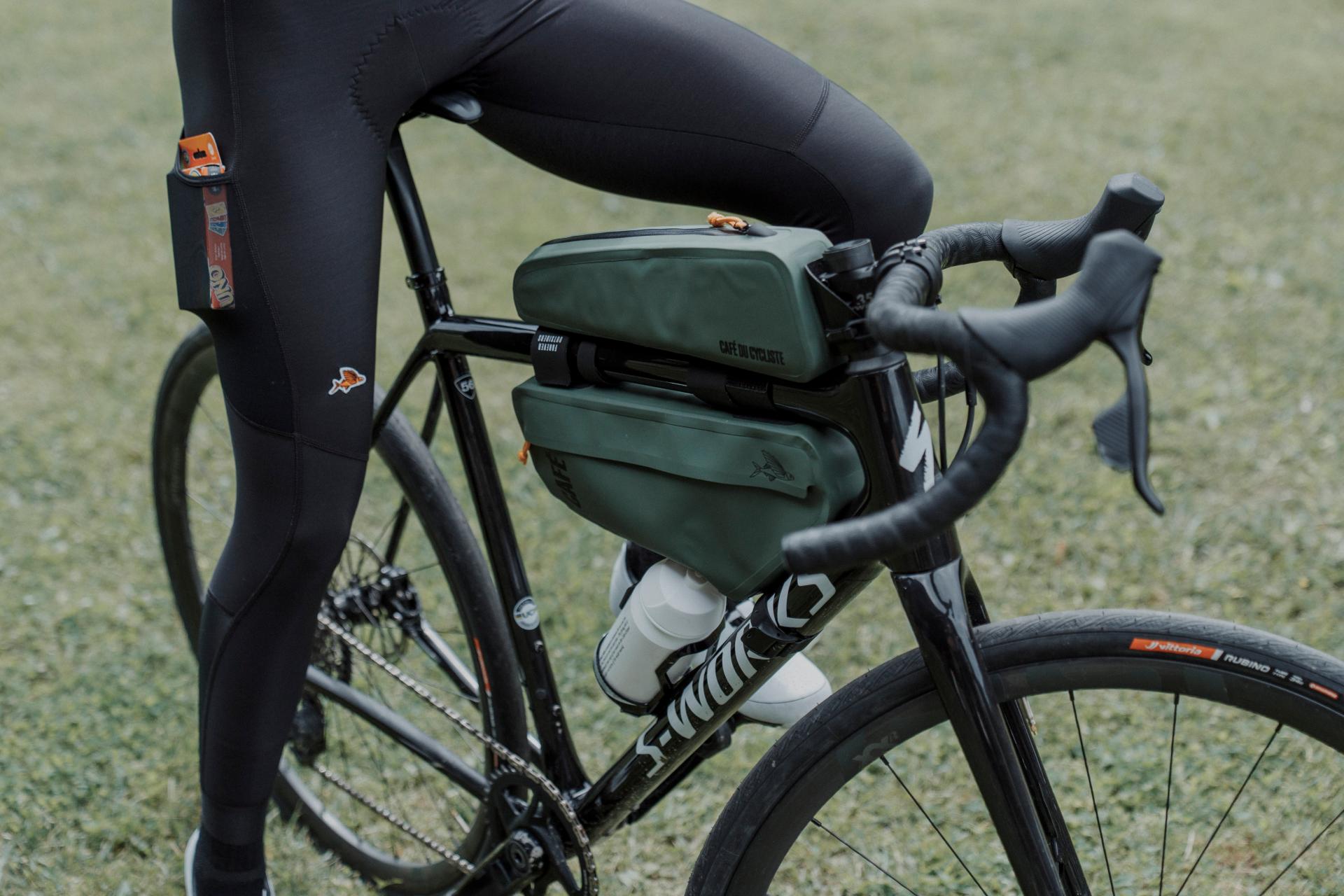 A close-up outdoor shot of a cyclist wearing black cycling tights and a black road bike, with an olive green frame bag mounted on the bicycle frame. The background is a soft-focus grassy field.