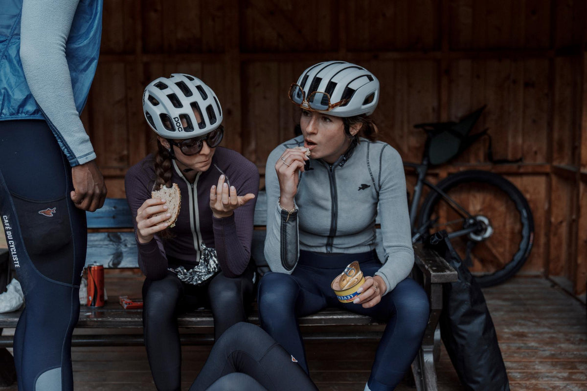 Two cyclists sitting on a wooden bench in a rustic indoor setting, wearing helmets and cycling jerseys while eating sandwiches. A bicycle is parked in the background.