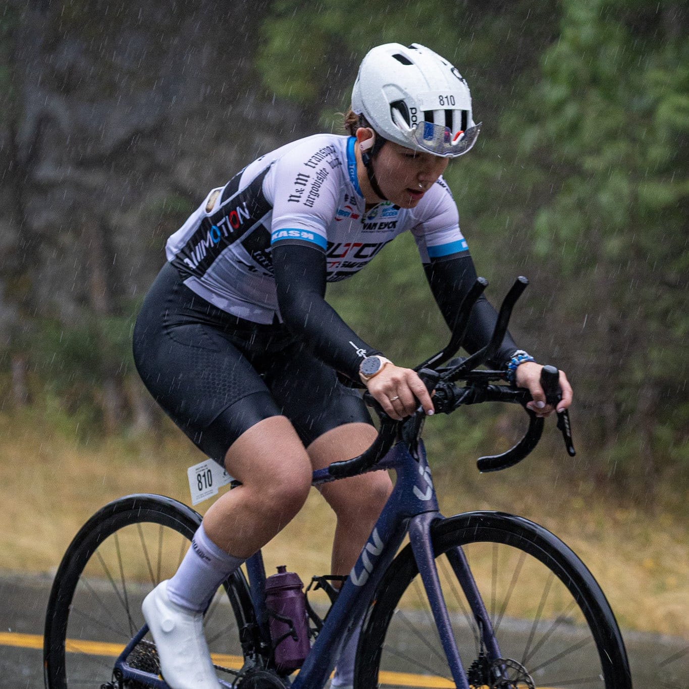 A cyclist in a white, black, and blue cycling jersey rides a dark blue road bike on an asphalt road with trees in the background.