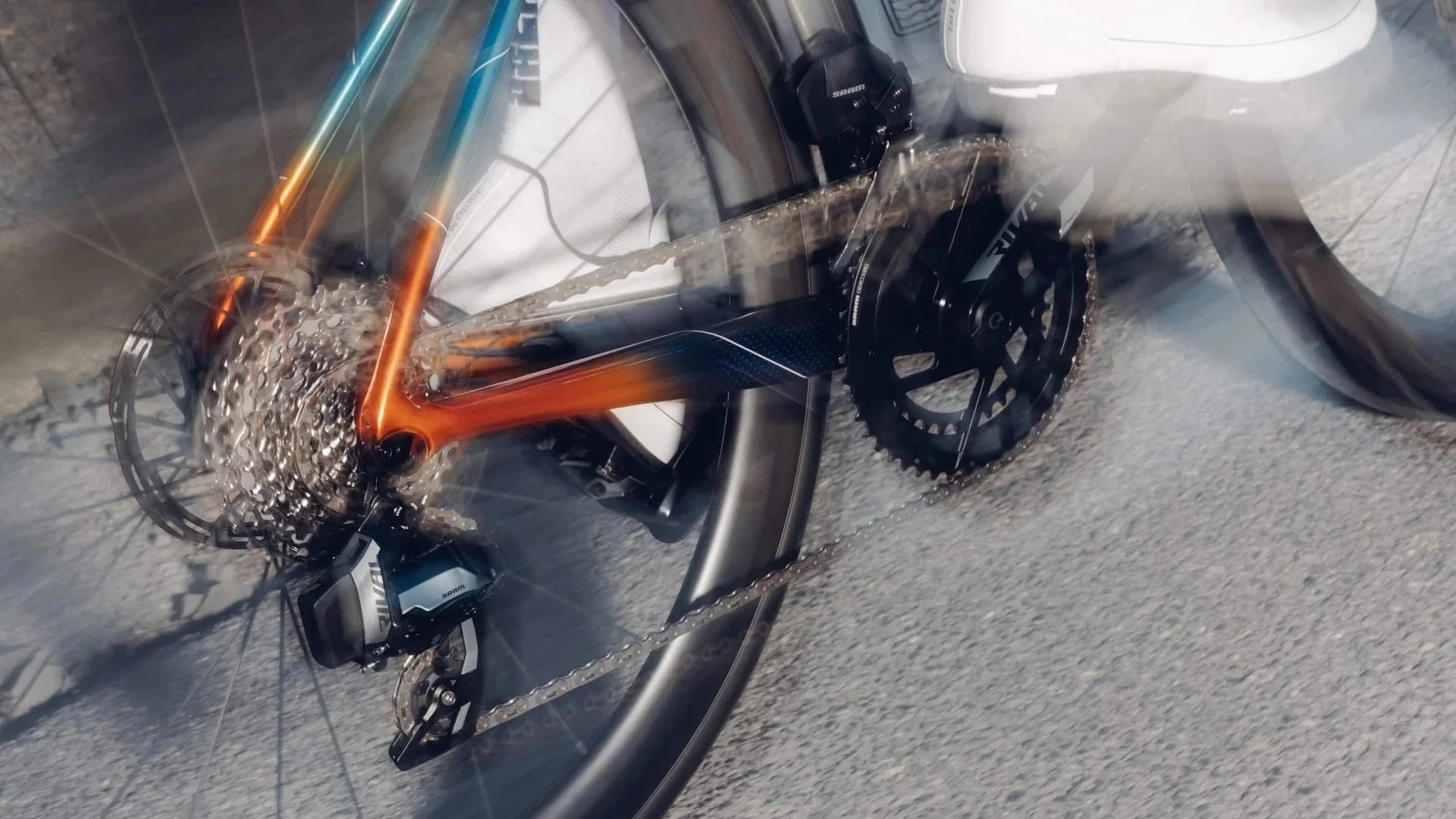 Close-up motion shot of a SRAM Rival crankset and rear derailleur in action on an orange and teal road bike, highlighting the drivetrain’s smooth performance and 2x gearing setup