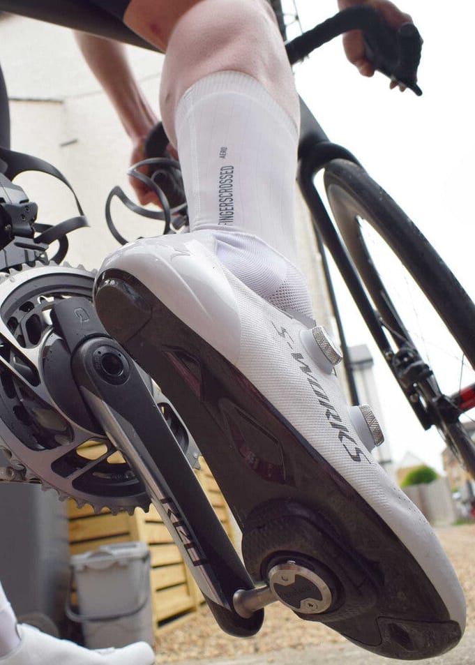 A close-up lifestyle shot of a cyclist's leg and foot on a road bike, showcasing a white and black Specialized cycling shoe with a white crew sock.