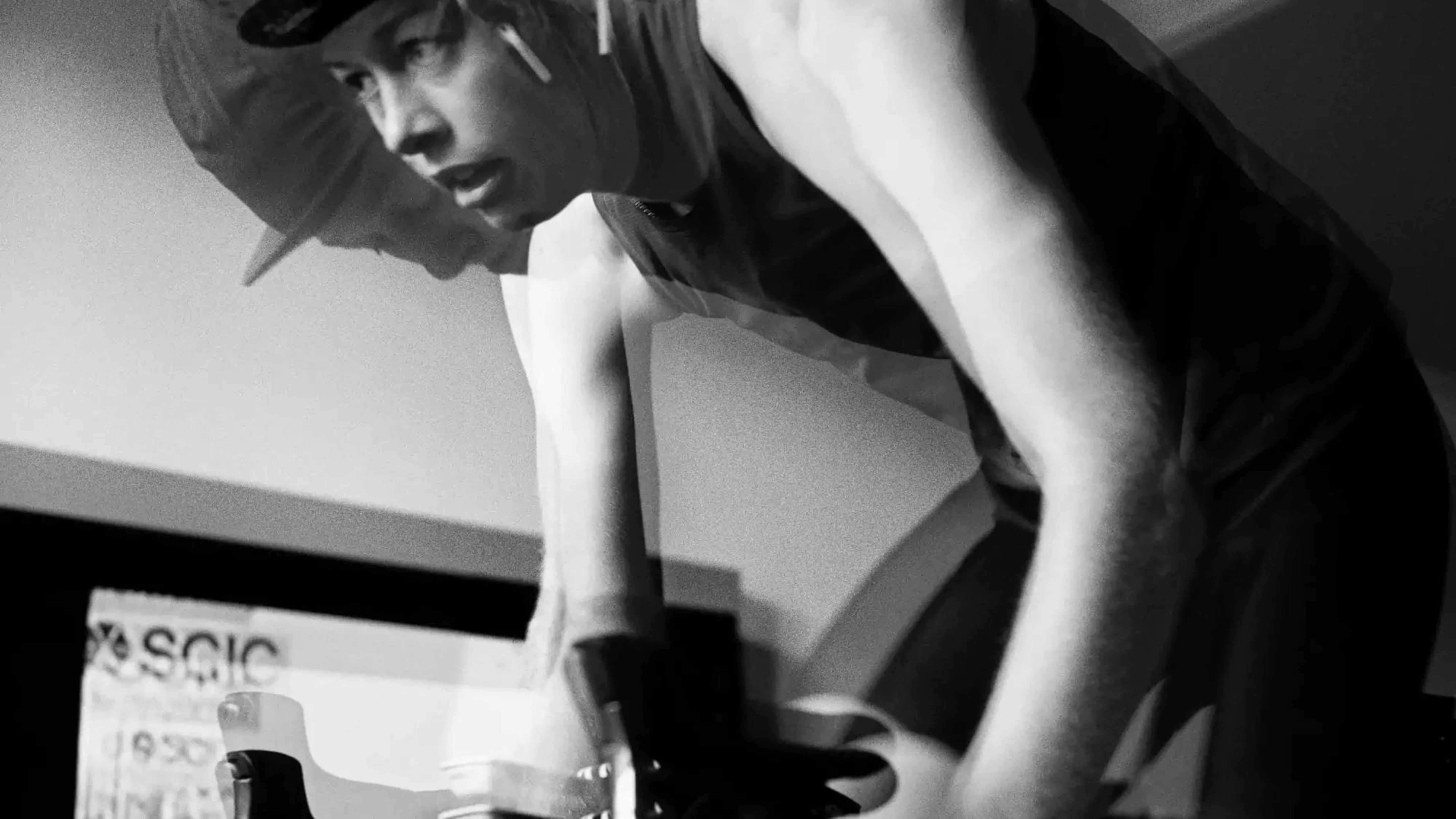 A black and white photograph of a cyclist hunched over the handlebars, gripping them tightly. A double exposure effect creates a ghostly duplicate of their face and upper body, emphasizing motion and exertion