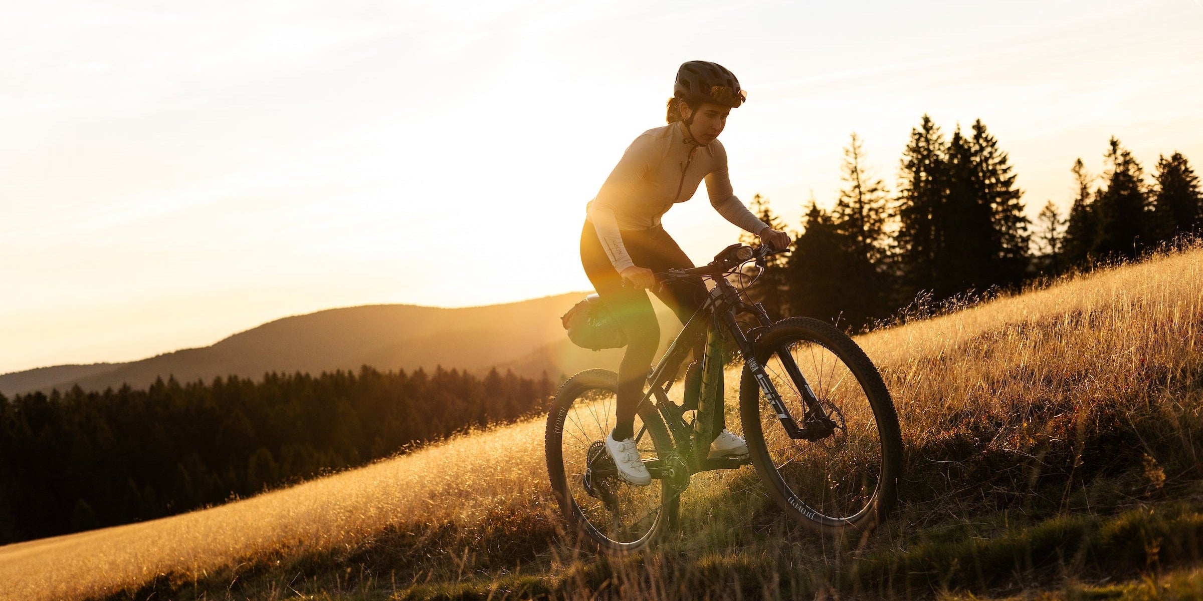 Person riding a bicycle on a grassy hill with trees and mountains in the background during sunset.