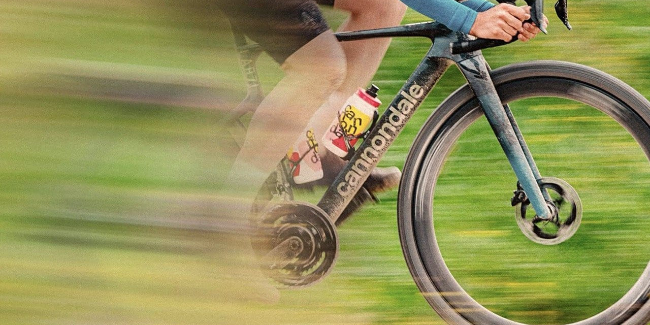 A close-up shot of two cyclists riding a Cannondale gravel bike at high speed. Motion blur surrounds the scene, emphasizing movement. One rider wears a blue long-sleeve jersey, gripping the handlebars, while the other’s legs are visible pedaling. A colorful water bottle with a playful design is attached to the frame.